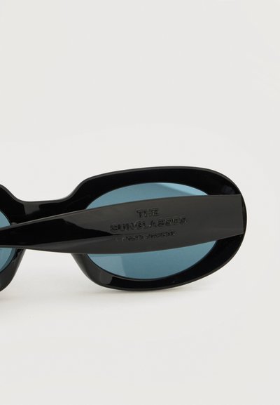 Μαύρα οβάλ γυαλιά ηλίου με σκουρό μπλε φακούς και εμφανές λογότυπο χαραγμένο στο πλαινό μέρος του μπράτσου, πάνω σε απλό λευκό φόντο.