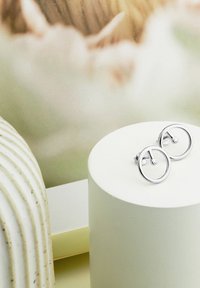 Silver cirkulära stiftörhängen med en liten metallisk stift i mitten, uppställda på ett runt vitt podium mot en mjuk, ljus bakgrund.