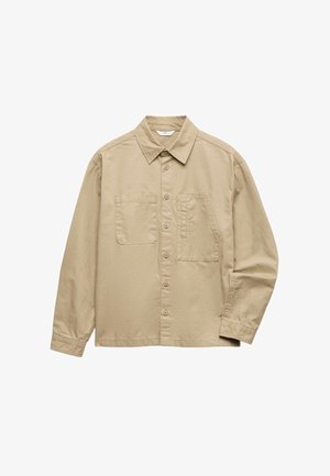 Beige Hemd mit Knöpfen, schwarzes T-Shirt darunter, dunkle Jeans und braune Sportschuhe. Lässiges Outfit, lockere Passform und hochgekrempelte Ärmel.