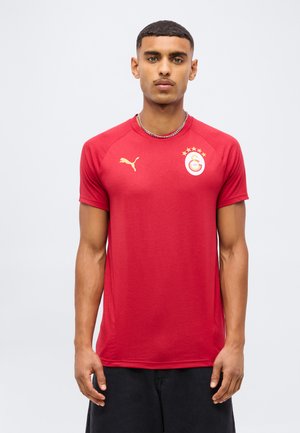Jeune homme portant un maillot de sport rouge Puma avec le logo du club de football Galatasaray, debout devant un fond clair uni.