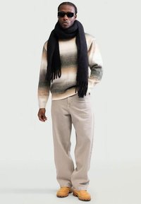 Pulover tricotat cu model de valuri în crem, verde și marron, asociat cu pantaloni bej deschis și saboți galbeni; o eșarfă neagră și ochelari de soare finalizează aspectul.