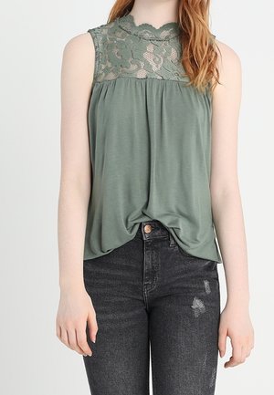 Mouwloos saliegroen topje met kanten inzetstuk, gecombineerd met hoog getailleerde zwarte distressed skinny jeans bij een staande persoon.