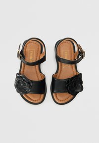 Sorte skinnsandaler med en bred stropp, justerbar ankelspenne og en blomsterdetalj på siden. Myk brun såle med stikking.