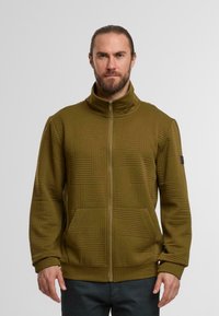Mann mit Bart und zurückgebundenem Haar trägt eine olivgrüne, gemusterte Zip-Jacke und dunkle Hosen vor einem schlichten hellen Hintergrund.