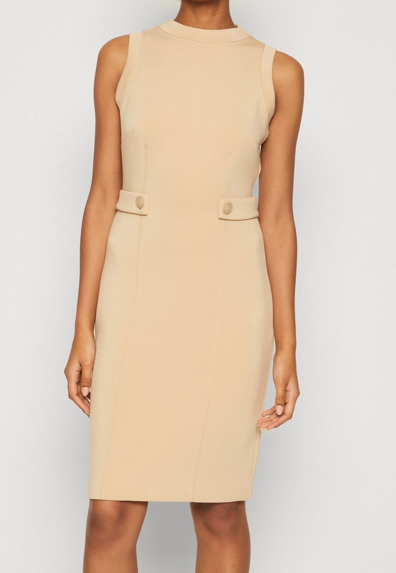 Robe beige sans manches longueur genou avec un col montant et des pattes latérales décoratives avec boutons, portée par une personne debout.