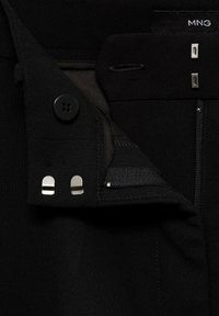 Gros plan sur la ceinture d'un pantalon noir montrant une agrafe et une barre, un bouton, une fermeture éclair et une partie d'une étiquette en tissu portant la mention MNG. Aucune personne visible.