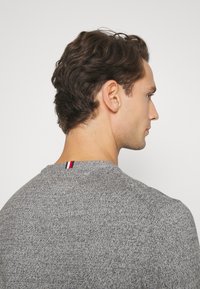 Pull en tricot gris avec un col rond, présentant une texture subtile et un accent rouge, blanc et bleu au niveau de la nuque arrière.