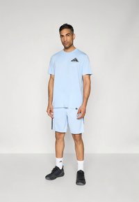 Lys blå T-skjorte med korte ermer og svart Adidas-logo på brystet, kombinert med matchende shorts med svarte sidelister, brukt sammen med svarte sko.