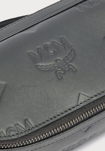 Κοντινή λήψη μαύρης δερμάτινης τσάντας με ανάγλυφο λογότυπο MCM και σχέδιο δάφνης, με φερμουάρ από ασημένιο μέταλλο.