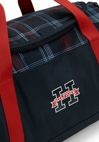 Borsa da viaggio in nylon nera con manici rossi, caratterizzata da un motivo scozzese rosso e blu. Include un logo "HILFIGER" ben visibile sul lato.