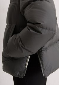 Zwarte gewatteerde jas met een mouw en zijkant, waarvan de rits gedeeltelijk open is, met het "Marco O'polo" label zichtbaar op een neutrale achtergrond.