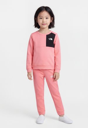 Jeune fille debout portant un sweat-shirt rose et un pantalon avec des écussons noirs, assortis à des chaussures blanches sans lacets sur fond blanc.