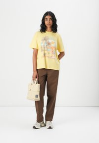 Gul grafisk t-shirt med palmträdsmönster, kombinerat med bruna byxor. Håller en beige tygväska med framfickor. Grå sneakers.