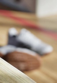 Chaussures de sport en focus, coloris gris et blanc, texture visible. Un ballon marron flou en arrière-plan sur une surface en bois.