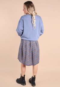 Sweatshirt bleu clair avec des épaulettes tombantes, associé à une jupe patineuse de longueur genou à motifs géométriques bleus, orange et marron. Bottines noires.