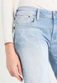 De jeans de mezclilla azul claro con un parche descolorido y un ligero desgaste en el muslo. Diseño clásico de cinco bolsillos con una cintura de altura media.