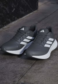 Paire de chaussures de course Adidas noir et blanc avec trois bandes blanches, posée sur une surface pavée en pierre.