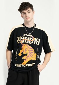 Młody mężczyzna z krótkimi włosami, ubrany w czarną i beżową koszulkę z kolorową grafiką tygrysa i napisem "NIE DO ZATRZYMANIA".