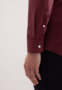 Linker Arm einer Person in einem kastanienbraunen Langarmshirt mit weißen Knöpfen und einer kleinen bestickten Rose am Bündchen, die Hand entspannt an der Seite vor einem hellen Hintergrund.