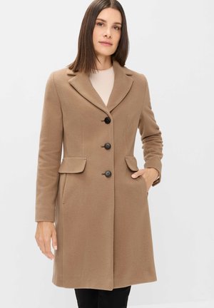 Woman wearing a fitted camel-brown coat with three black buttons, notch collar, flap pockets, and one hand in pocket, standing against white background.