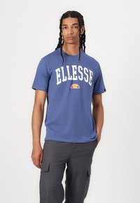 Mėlynas medvilninis marškinėliai su balta "ELLESSE" logotipu, virš teksto yra daugybės spalvų akcentas. Kasdienė laisvo kirpimo su trumpomis rankovėmis.