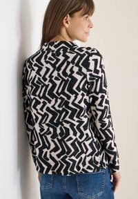 Schwarz-weiß gemusterten Pullover mit Zickzackdesign, leichtem Stoff, rundem Ausschnitt, langen Ärmeln und seitlichen Schlitzen, kombiniert mit blauen Jeans.