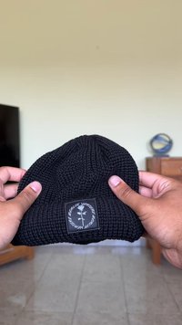 Gorro de malha preto com um padrão texturizado e um patch retangular com texto branco e design gráfico. Aba dobrada, material suave.