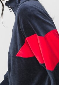 Marineblauwe hoodie met rode veloursaccenten op de mouwen. Voorzien van een hoge kraag en ritssluiting, een zachte textuur en een zak aan de voorkant.