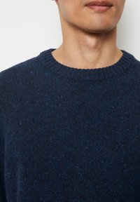 Dunkelblauer Pullover mit einem gerippten Rundhalsausschnitt. Die Textur ist gestrickt und weist Punkte in helleren Blautönen und dunkleren Nuancen auf.