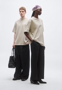 Tricouri supradimensionate bej, pantaloni negri largi și pantofi negri. Modelul din stânga ține o geantă tote neagră; modelul din dreapta poartă un batic violet cu model.