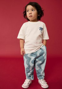 Fehér póló, a mellkasán pálmafás mintával, kék hullámos mintás nadrággal és tépőzáras fehér tornacipővel párosítva. A háttér piros.