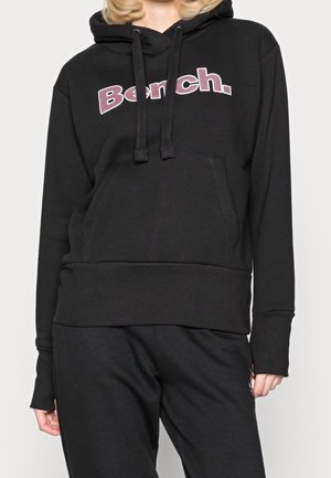 Person trägt einen schwarzen Bench-Hoodie mit Fronttasche und Kordelzug, kombiniert mit schwarzen Hosen vor einem einfarbigen hellen Hintergrund.