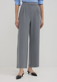 Graue, maßgeschneiderte, weit geschnittene Hosen mit Falten und einem seitlichen Knopfdetail, aus einem glatten Material, kombiniert mit schwarzen Slingback-Pumps.