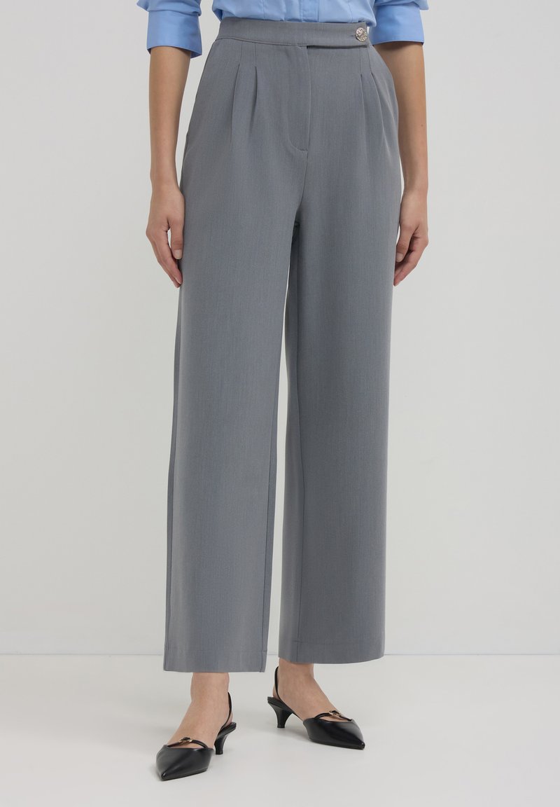 Graue, maßgeschneiderte, weit geschnittene Hosen mit Falten und einem seitlichen Knopfdetail, aus einem glatten Material, kombiniert mit schwarzen Slingback-Pumps.