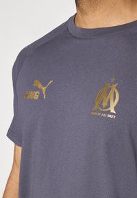 Mørkeblå T-shirt med gyldent "KING" tekst og Puma-logo på venstre bryst, samt et Marseille-emblem og "DROIT AU BUT" nedenfor til højre.