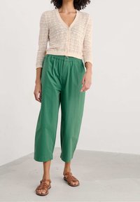 Cárdigan de punto en beige claro con patrón de tejido abierto, combinado con pantalones verdes holgados y sandalias planas marrones con diseño de tiras.