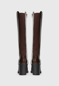 Ett par långa, mörkbruna läderboots med klack och elastiska paneler, sedd bakifrån mot en enkel ljus bakgrund.