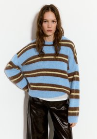 Pulover cu dungi albastre, cu dungi maro și crem, realizat dintr-o textură pufoasă, purtat cu pantaloni largi negri lucioși.