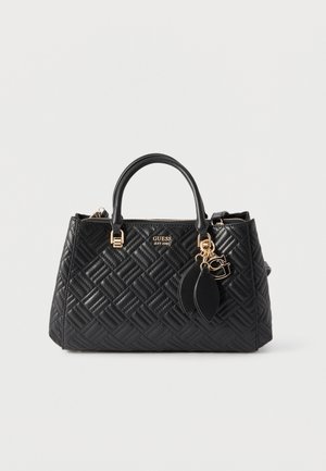 Sac à main en cuir noir matelassé avec doubles poignées, accents dorés, pendentif décoratif et logo "GUESS EST 1981" sur le devant.