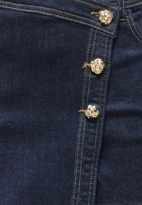 Tissu en denim bleu foncé avec une rangée verticale de trois boutons décoratifs en forme de tête de lion dorée et un détail de couture beige.