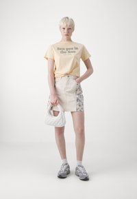 Kremfarget denim mini-skjørt med blomstrete sidelapper, kombinert med en gul t-skjorte med grafikk. Tilbehør inkluderer en hvit quiltet håndveske.