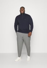 Sweat à zip bleu marine avec des poignets côtelés, associé à un pantalon rayé gris et blanc. Des baskets blanches complètent la tenue.