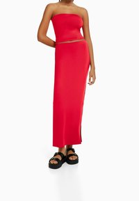 Czerwona bluzka bez ramiączek połączona z dopasowaną długą spódnicą z bocznym rozcięciem, wykonaną z gładkiego materiału, noszona z czarnymi sandałami.