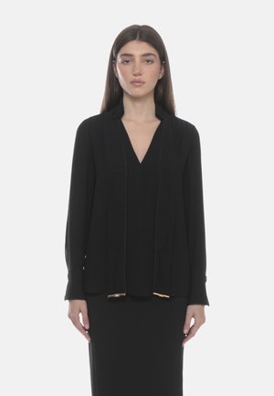 Mujer con cabello castaño liso, vestida con una blusa negra de escote en V y un blazer negro, posando frente a un fondo claro y liso.