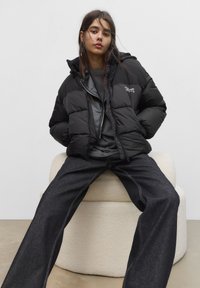 Giacca nera imbottita con cappuccio, zip frontale e logo ricamato; indossata sopra una maglietta grigia e abbinata a jeans in denim scuri.
