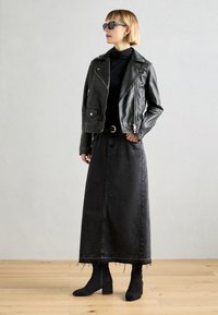 Veste de moto en cuir noir sur un col roulé noir, associée à une longue jupe en denim noire et des bottines noires à talons texturés.
