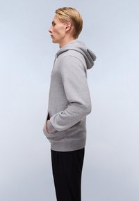 Grijze katoenen hoodie met een voorzak, verstelbare trekkoordcapuchon, ribgebreide manchetten en zoom, gedragen over zwarte broeken. Zijaanzicht.