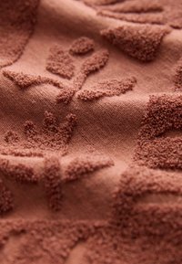 Tissu texturé de couleur rouille présentant des motifs floraux en relief. La surface affiche une qualité douce et pelucheuse avec des détails complexes.