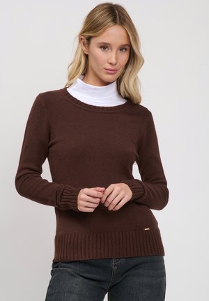 Jonge vrouw met blond haar die een bruine trui draagt over een witte coltrui en donkere spijkerbroek, staand tegen een lichte achtergrond.