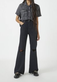 Henkilö, jolla on päällä mustat leveät farkut, joissa on polvireiät, musta napitettava lyhythihainen paita ja mustat loafer-kengät, joissa on kultaketjuja.